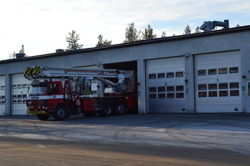 Bilden visar ett brandfordon, en röd Scania räddningsbil med lyftplattform, som delvis är parkerad vid en brandstation. Fordonet står framför en av byggnadens portar och lyftplattformen är delvis utkörd ur porten. Brandstationen är en grå byggnad med