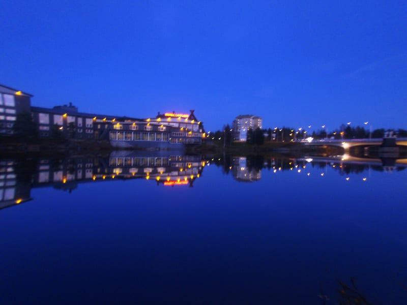 En bild tagen i skymning eller natt som visar en byggnad med upplysta fönster och en skylt som reflekteras i ett mörkt vatten. Byggnaden är belägen vid vattnet, med träd och buskar som omger den. Längre bort syns en bro med gatlyktor som också reflek