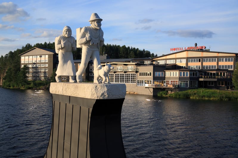Bilden visar ett landskap med en sjö eller flod i förgrunden och byggnader samt skog i bakgrunden. En skulptur föreställande en kvinna, en man med ryggsäck och en hund står på en piedestal nära vattnet. Mannens hatt och kläder samt ryggsäcken kan ind