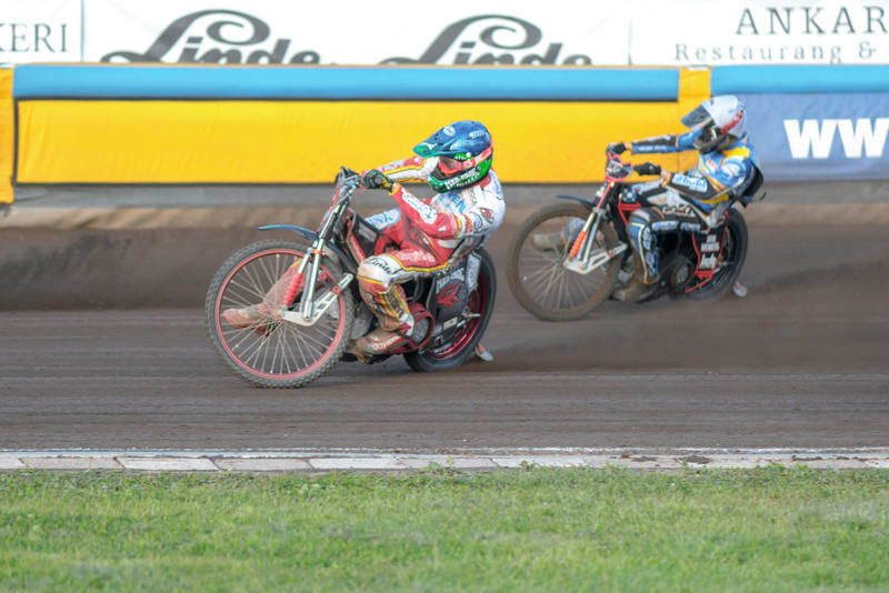 Bilden visar två speedwayförare på en grusbana i full fart. Föraren i förgrunden bär röd, vit och svart racingdräkt med grön hjälm, och hans motorcykel har röda hjul. Bakom honom syns en annan förare i blå och vit racingdräkt med en vit hjälm, och de