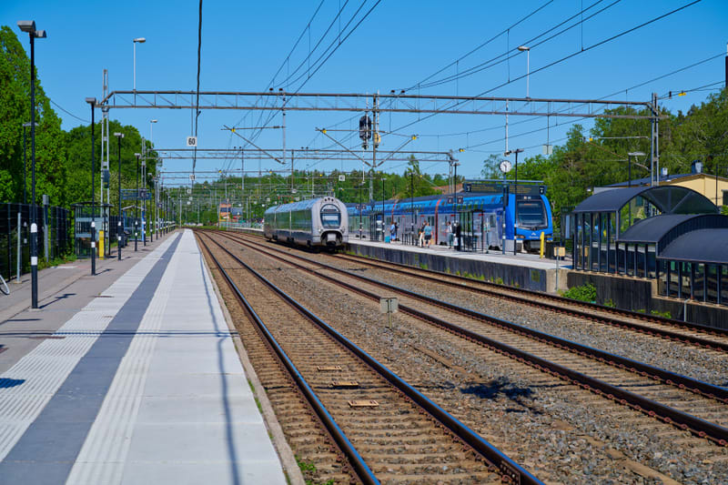 Bilden visar ett tågstopp eller en järnvägsstation utomhus under dagtid med klarblå himmel. Ett silverfärgat tåg står på ett av spåren i mitten av bilden, och ett blått tåg med gula detaljer står vid en perrong till höger. Flera personer syns på perr