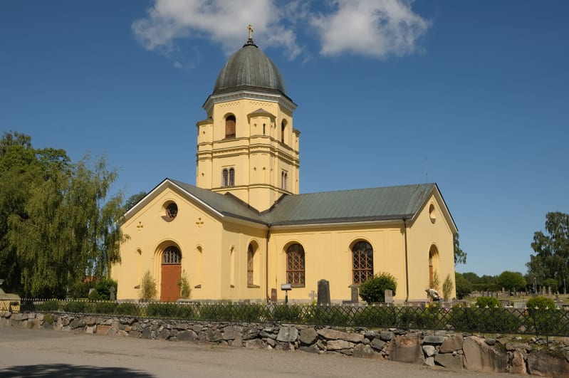 En gul kyrkobyggnad avbildad från sidan i soligt väder med en klarblå himmel i bakgrunden. Kyrkan har en stor rundad kupol på sitt klocktorn, som är delvis synligt. Byggnaden har flera fönster med runda överdelar och träramar. Ett brungrått tak täcke