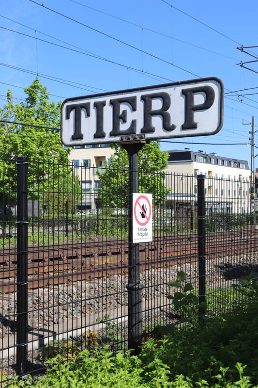 Bilden visar en järnvägsskylt med texten ”TIERP” i svart på vit bakgrund, monterad på en stolpe. Under skylten sitter en mindre rund skylt med ett rött förbudsmärke som visar en hand med ett rött streck över, samt texten ”Tillträde förbjudet”. I förg