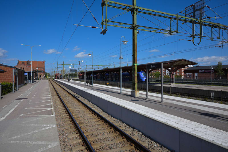 En bild tagen i dagsljus som visar en järnvägsstation från spårområdet. I förgrunden syns tågspår med grus mellan rälsen. Till vänster om spåren finns en plattform med zebramönstrade markeringar och en byggnad i rött tegel. Längre bort på plattformen