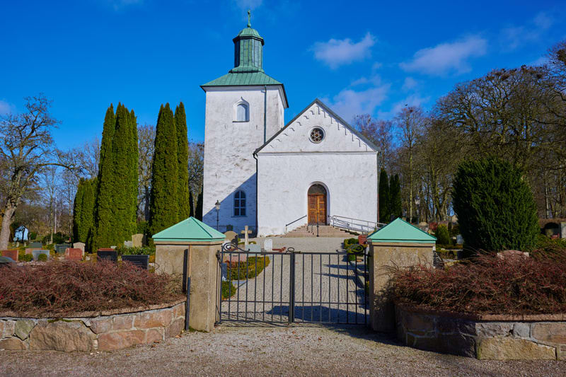 Bilden visar en vit kyrkobyggnad med ett grönt tak och en klockstapel, centralt placerad. Intill kyrkan finns en kyrkogård med gravstenar och träd, varav några är kala medan andra är barrträd med gröna barr. Framför kyrkan syns en smidesgrind med två