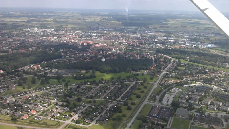 En vidvinkelbild tagen från ovan, troligen från ett flygplan, visar ett bebyggt område omgivet av grönområden och åkrar. I förgrunden syns ett bostadsområde med enfamiljshus och trädgårdar, samt ett område med flerbostadshus och parkeringsplatser. Et