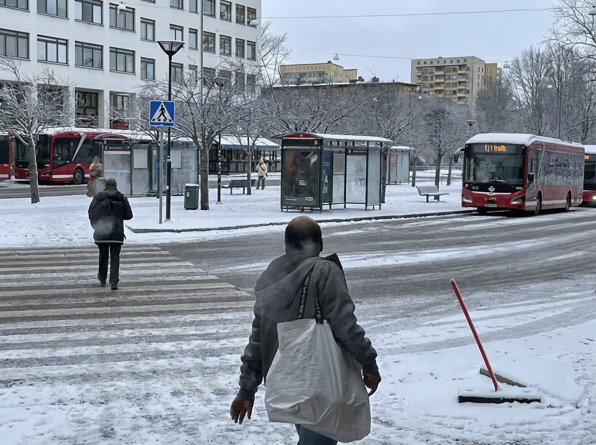 En ganska hög bild tagen med en nedåtvinklad position, med betraktaren placerad bakom ryggen på en person av obestämd ålder och kön som går mot en väg. På vägen till höger syns en röd buss med texten "EJ I TRAFIK" på displayen, och det verkar finnas