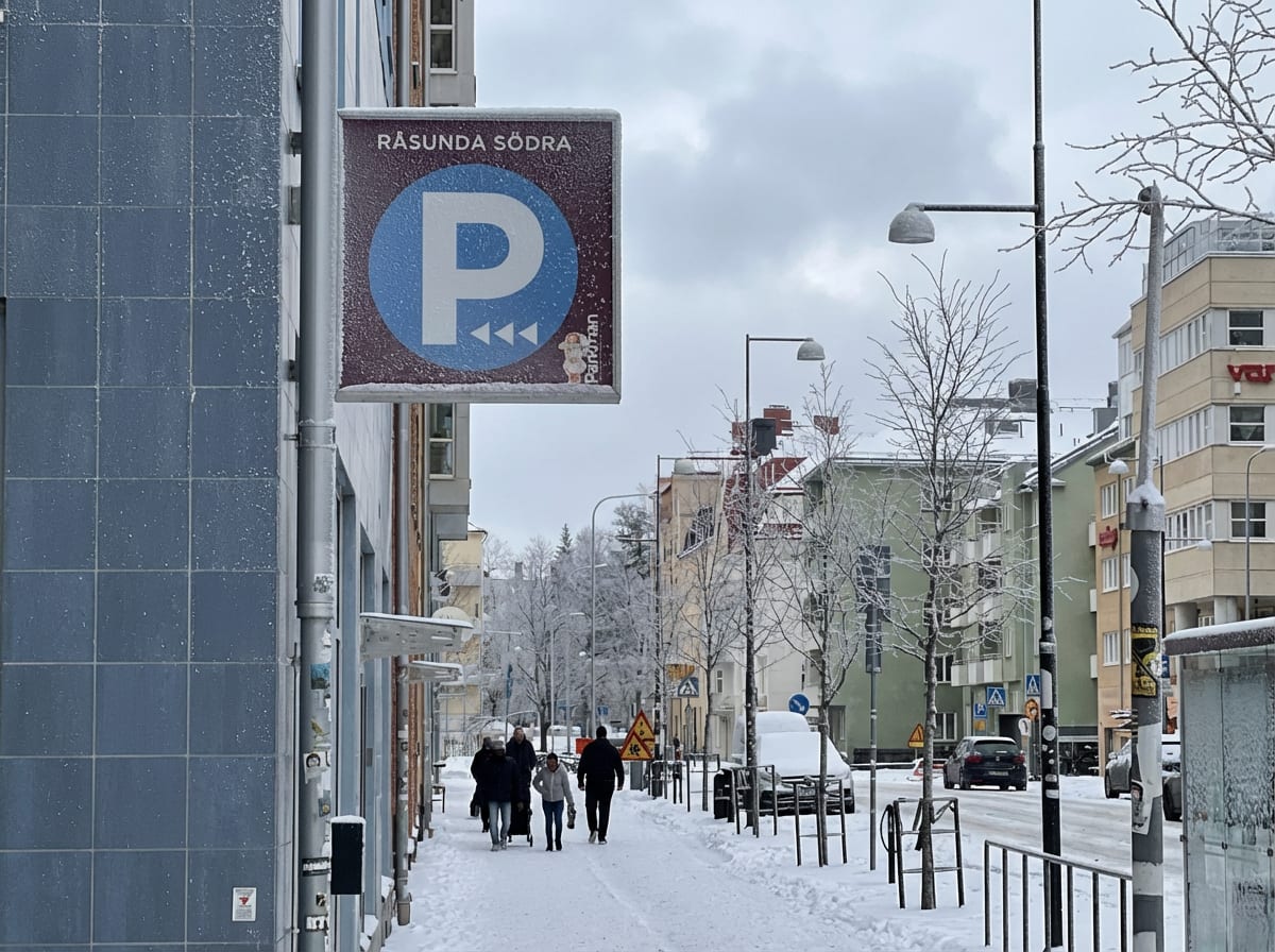 En ganska bred bild av en gata under vintertid. Till vänster i förgrunden syns en blå kaklad vägg och en stolpe med en stor skylt med texten "RÅSUNDA SÖDRA" och en blå P-symbol. Skylten ser ut att vara täckt av is eller frost. Gatorna och träden är t