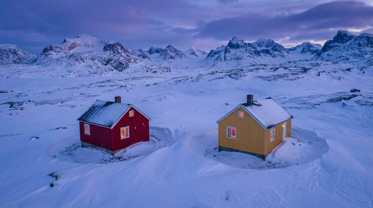 Bilden visar ett snötäckt, bergigt landskap under en molnig himmel i skymningen eller gryningen. I förgrunden står två små, fristående hus. Det vänstra huset är rött med vit dörr och flera fönster som lyser varmt inifrån. Taket är snötäckt och det ha