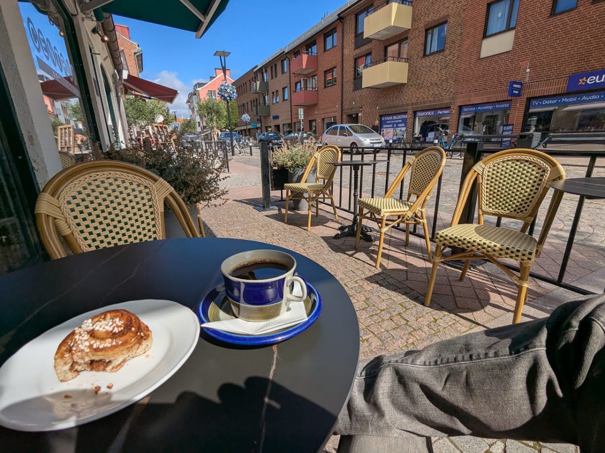 Bilden visar ett gatuplan med en fikabord och omgivning utomhus i soligt väder. I förgrunden syns ett svart runt bord med en biten kanelbulle på ett vitt fat och en kaffekopp på ett fat. En del av en persons ben i mörka byxor syns i nederkanten av bilden. Bortom bordet sträcker sig en kullerstensbelagd mark med flera tomma rotinklädda caféstolar runt svarta bord. En mörk grind eller avspärrning går längs kanten av trottoaren. I bakgrunden syns en byggnad med rött tegel, fönster och balkonger. På byggnadens bottenvåning finns butiksfönster med skyltar, bland annat en med texten "Euronics". En ljus bil och cyklar är parkerade längs gatan. Växter i krukor och hängande dekorationer bidrar till miljön. Himlen är klarblå med några vita moln.