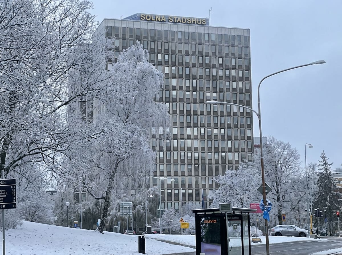 Bilden visar Solna Stadshus en vinterdag med snö och rimfrost. En stor byggnad med många fönster dominerar mitten och höger sida av bilden. Ovanpå byggnaden syns texten "SOLNA STADSHUS" i guldfärgade bokstäver. Framför byggnaden, till vänster och i f