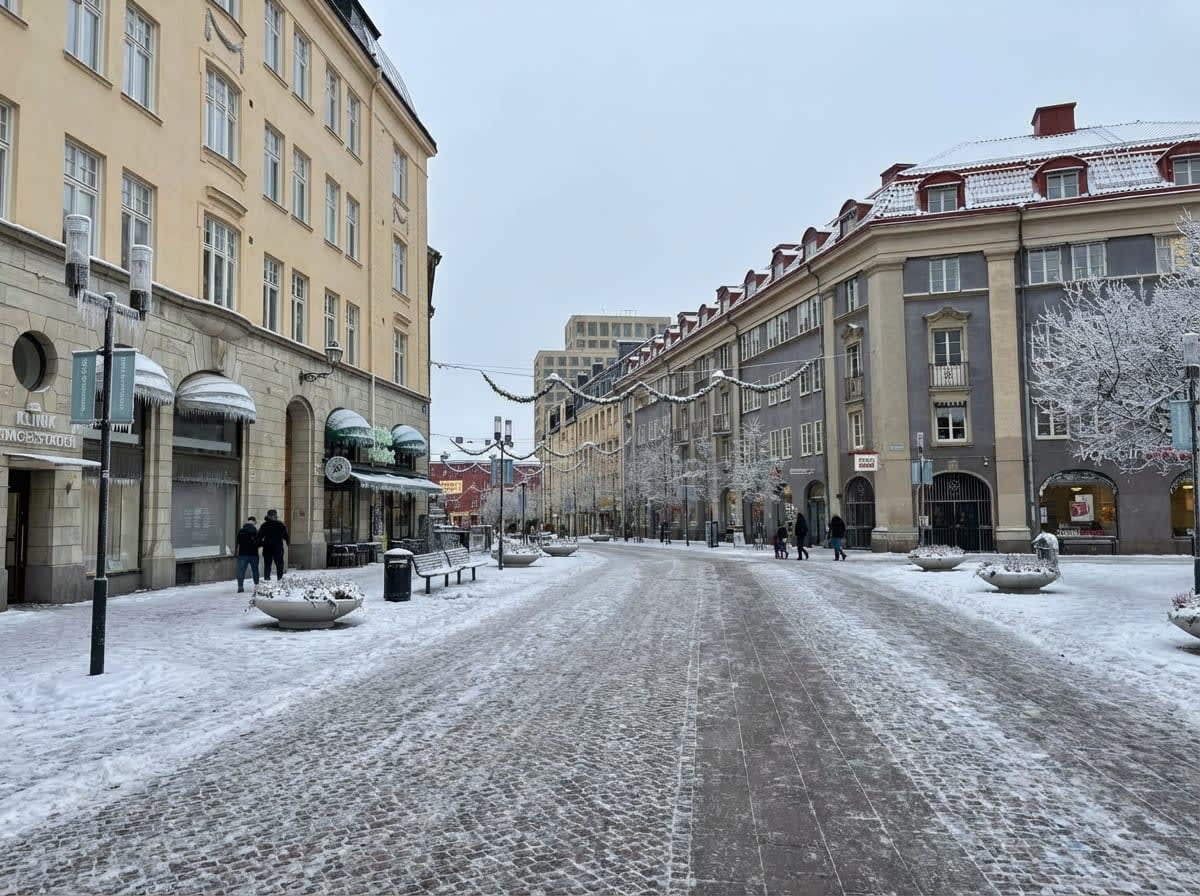 Bilden visar en snötäckt stadsgata kantad av byggnader under dagtid. Gatan är bred och belagd med gatsten, delvis täckt av snö och is. På båda sidor av gatan syns flervåningsbyggnader, den till vänster är ljusgul medan den till höger är gråbrun med röda tak. Fönstren är vita och har rutor. På gatan finns bänkar, soptunnor och runda planteringar som också är täckta av snö. Mellan byggnaderna hänger ljusslingor, sannolikt juldekorationer. Träd längs gatan är täckta av rimfrost eller snö. Ett fåtal personer promenerar på trottoarerna, klädda i varma kläder. Himmeln är ljus och molnig, vilket indikerar att det är dagsljus men inte soligt. Vid en av byggnaderna finns skyltar som delvis syns, bland annat en med texten 'KLINIK'. Byggnaderna har markiser, varav några är snötäckta. Flera istappar hänger från takfoten på byggnaden till vänster.