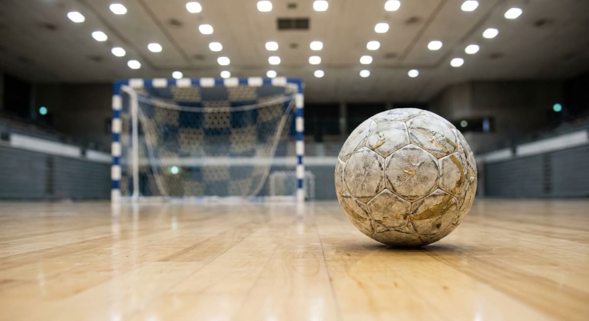 Bilden visar en sliten handboll i fokus på en glänsande trägolv i en idrottshall. Bollens yta är vitaktig med mörkare partier och strukturen av sömmarna syns tydligt. I bakgrunden, något ur fokus, står ett handbollsmål med blå- och vitrandiga stolpar och ett vitt nät. Taket i idrottshallen har en serie tända taklampor, som reflekteras i golvet. Längre bak syns sittplatser, i form av läktare. Ljuset i förgrunden är starkare än i bakgrunden.