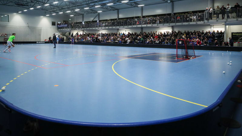 En inomhusbild som visar en stor, ljusblå idrottsplan, troligen för innebandy. En person klädd i grön tröja och vita byxor håller en innebandyklubba och syns på vänster sida av bilden. Planen har vita, orangea, gula och röda linjer samt ett mörkt blå