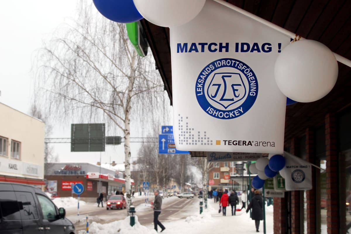 Bilden visar ett gatuplan täckt av snö från ett butiksportskontor, troligen i Leksand, Sverige. En skylt med texten 'MATCH IDAG' och 'Leksands Idrottsförening Ishockey' samt 'Tegera arena' syns tydligt i förgrunden, omgärdad av blå, vita och gröna ba