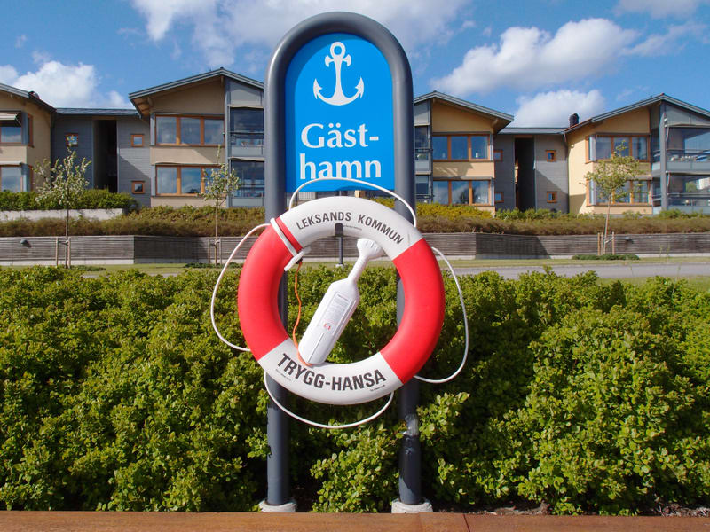 En bild tagen utomhus under dagtid visar en skylt för en gästhamn med en livboj hängandes på stolpen. Skylten är blå med en vit ankarsymbol och texten "Gästhamn". Livbojen är röd och vit med texterna "LEKSANDS KOMMUN" och "TRYGG-HANSA" tryckta på den