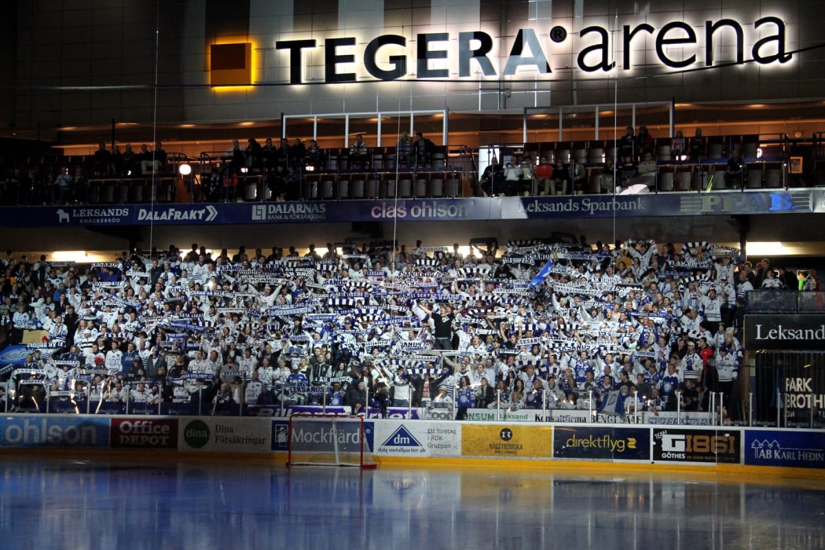 En stor publik av hockeyfans är samlad på läktaren i en ishockeyarena. Största delen av publiken är klädd i vita, blå och svarta färger och många håller upp halsdukar med texten "Leksands". Läktarna är fyllda med människor som ser ut att heja fram si