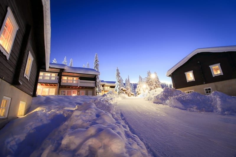 Bilden visar ett snötäckt landskap under vad som ser ut att vara skymning eller gryning, med en klar blå himmel som gradvis ljusnar mot horisonten. Flera byggnader med upplysta fönster syns. Till vänster finns en mörkare träbyggnad och längre bort i