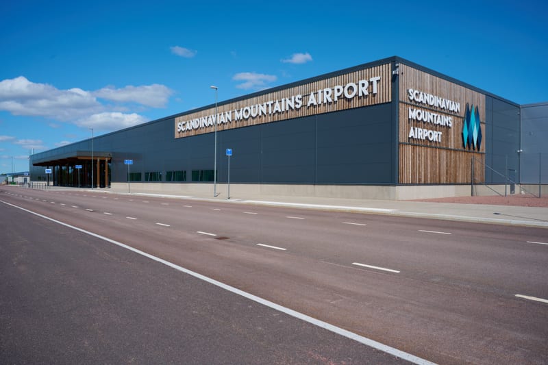 Bilden visar utsidan av en modern byggnad, identifierad med skyltar som "SCANDINAVIAN MOUNTAINS AIRPORT". Byggnaden är grå med stora fönsterpartier och delar klädda med vertikala träpaneler. Inför byggnaden finns en asfalterad väg med vita markeringa