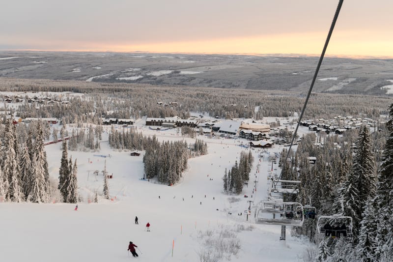 Bilden visar ett snötäckt landskap med en skidbacke och en by nedanför. I förgrunden syns flera skidåkare på en pistsnöig backe, och en del av en sittlift med flera sittplatser, vissa med passagerare. Liften sträcker sig uppåt, delvis utanför bildens