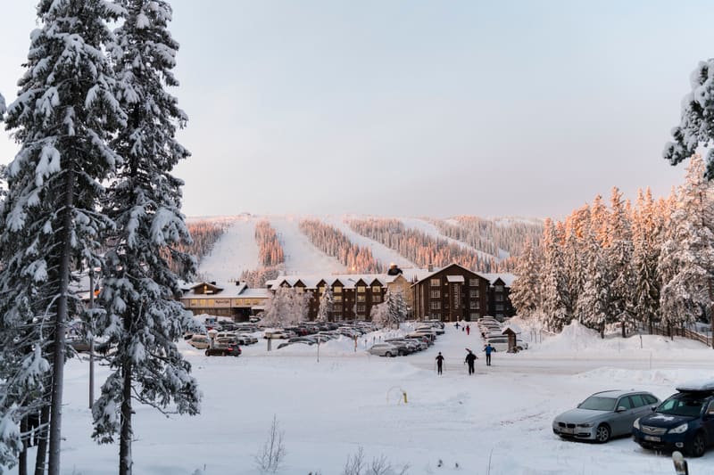 En vinterbild tagen utomhus som visar en skidort med flera byggnader omgivna av snöklädda träd. I förgrunden till vänster syns snötäckta granskogsträd. Mellan träden och byggnaderna finns flera parkerade bilar på en snötäckt parkeringsplats. På marke