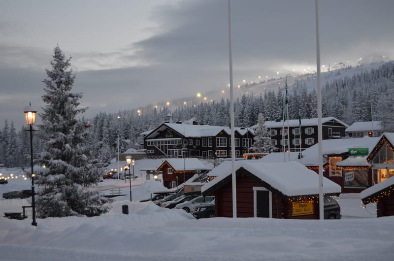Bilden visar ett snötäckt vinterlandskap med byggnader, träd och en skidbacke i bakgrunden. I förgrunden syns snötäckta träd, varav ett är dekorerat med julgranskulor. En gatlykta med lysande sken står till vänster i bilden. Flera byggnader i variera