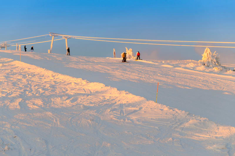 En bild tagen utomhus under dagtid som visar en snötäckt skidbacke under en klarblå himmel. Ljuset på snön har en varm, rosa-orange ton, vilket indikerar att det kan vara soluppgång eller solnedgång. Flera personer, iklädda vinterkläder, syns fördela