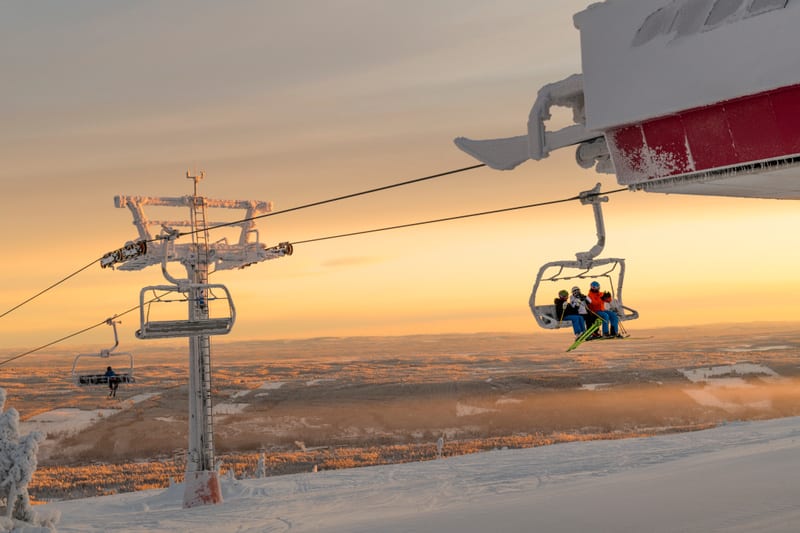 Bilden visar en skidlift i ett snötäckt, bergigt landskap under en solnedgång eller soluppgång. I mitten av bilden finns en stor liftstolpe täckt av snö och is. Till höger om stolpen är en liftkassett med tre personer, troligen skidåkare eller snowbo