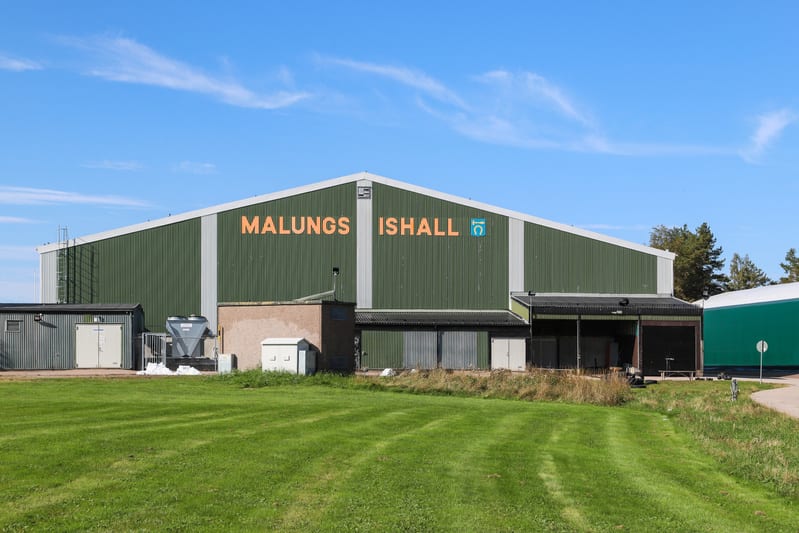 Bilden visar fasaden på en stor grön industriell byggnad under en solig dag. På byggnadens framsida står texten "MALUNGS ISHALL" i orange färg med en blå logotyp i mitten av texten. Nedanför huvudbyggnaden finns flera mindre byggnader och strukturer,