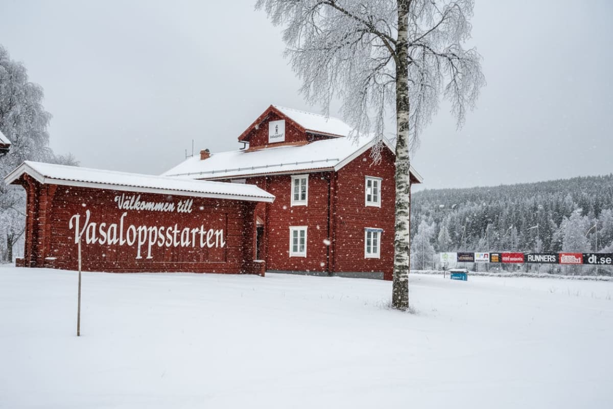 En vinterbild tagen under snöfall, med en röd träbyggnad till vänster i förgrunden och ett stort rött träskylt med texten "Välkommen till Vasaloppsstarten" framför byggnaden. Byggnaden har vita fönster och snötäckta tak. Till höger om byggnaden står ett snöigt lövträd med bar kvist. I bakgrunden syns en snötäckt skog och en rad med reklamskyltar på banderoller. Marken är helt täckt av snö. Snöflingor syns i luften.