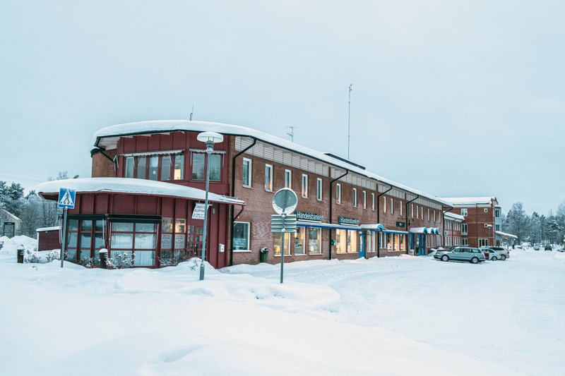 En bild tagen utomhus under vintertid visar en tegelbyggnad med ett rött tak och snö på taklisterna. Byggnaden inrymmer flera verksamheter, bland annat en Handelsbanken-filial med tydligt skyltade fönster. Framför byggnaden finns ett snötäckt parkeri