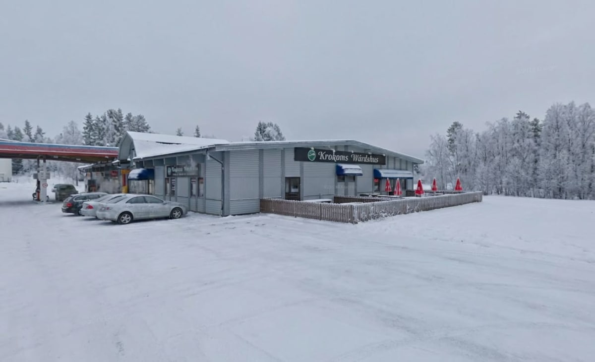 En yttre vy av en byggnad en snöig dag. Byggnaden har fasader i ljusgrått och ett tak täckt med snö. På byggnadens framsida finns skyltar, varav en större skylt lyder "Krokoms Wärdshus". Flera fönster och dörrar syns. Framför en del av byggnaden finns en terrass med ett brunt trästaket och flera (minst sex) röda parasoller hopfällda. En vit bil syns parkerad nära byggnaden. Till vänster om byggnaden finns en bensinstation med tak i rött och blått, samt bränslepumpar. I bakgrunden syns snötäckta träd och marken är helt täckt av ett tjockt snötäcke. Himlen är grå och mulen.