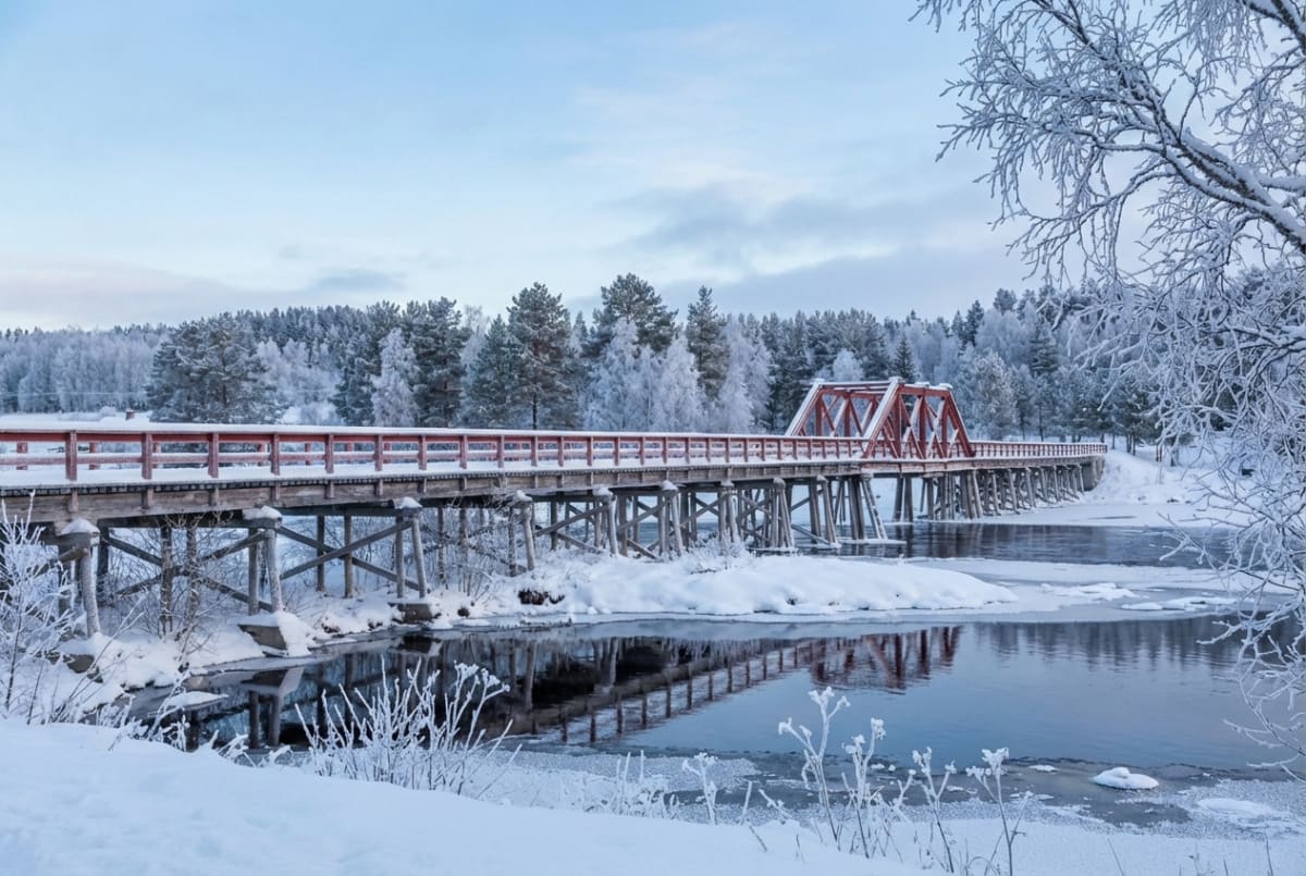 Bilden visar ett snöigt vinterlandskap med en bro som sträcker sig över en delvis frusen älv eller flod. Bron har en träkonstruktion med röda räcken, och i mitten finns en röd fackverkskonstruktion i metall. Träd och buskar som är täckta av snö och frost syns längs älvbankarna och i bakgrunden. Vattnet under bron reflekterar den ljusa himlen och delar av bron. Stranden och landskapet är helt täckt av snö. Himlen är ljusblå med inslag av tunna vita moln.