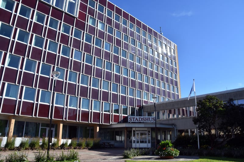 Bilden visar fasaden på en stor byggnad med många fönster. Byggnaden har en mörkröd fasad med vita ramar runt varje fönster. Fönstren är rektangulära och sitter tätt intill varandra i ett rutmönster. Framför byggnaden syns en ingång med en skylt ovan