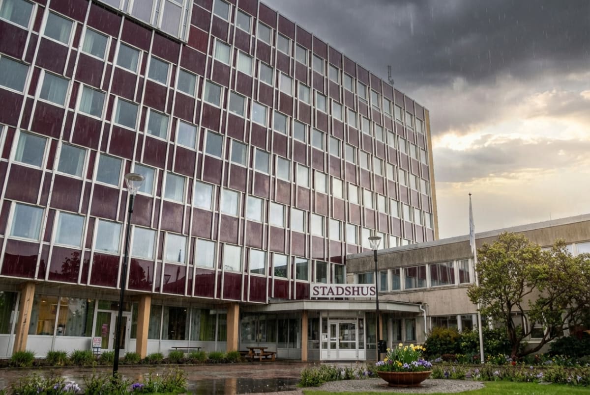 En bild av en stor byggnad med texten "STADSHUS" ovanför entrén. Byggnaden har många fönster och en fasad i mörkröda och ljusa färger. Nedanför byggnaden finns ett grönområde med blommor, buskar och träd. En stor blomkruka med färgglada blommor står på marken framför entrén. Marken framför byggnaden är fuktig, vilket indikerar regn, och det syns tydligt regndroppar i luften mot en mörk och molnig himmel. En flaggstång med en vit flagga märkt med en symbol syns till höger om byggnaden.