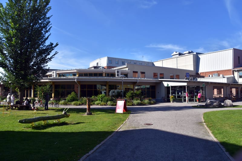 En utomhusbild som visar en modern byggnad med stora fönster och en entré. Framför byggnaden finns en gräsmatta med ett par lekredskap, inklusive en gungbräda formad som en grön larv och andra mindre, färgstarka föremål. En grusgång leder fram till e