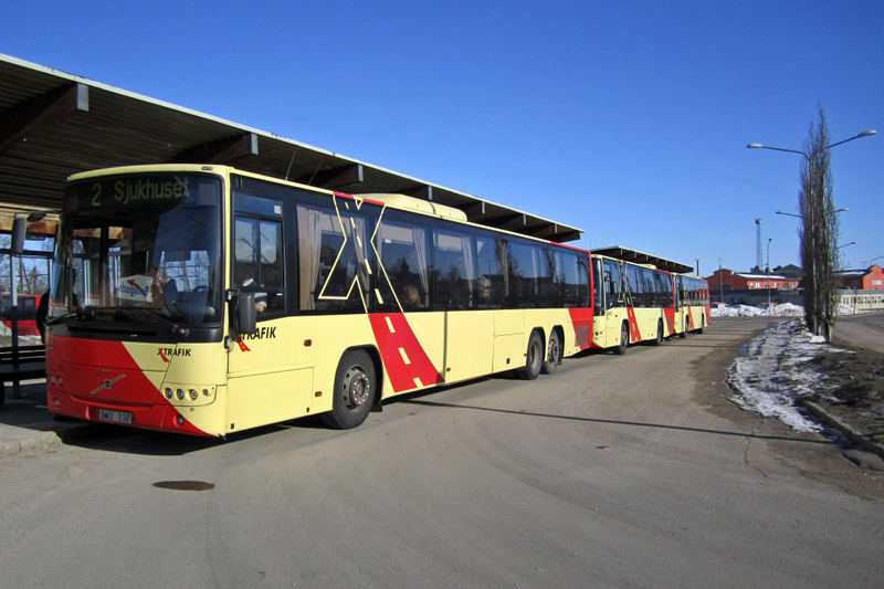 En gul och röd ledbuss av märket Volvo står parkerad vid en busshållplats under en klarblå himmel. Bussens front är röd och gul, och på sidan har den ett rött mönster med texten "XTRAFIK". Ett tydligt destinationsmeddelande på bussens front anger "2