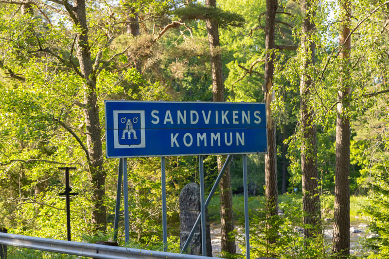 En blå skylt för 'SANDVIKENS KOMMUN' står i fokus framför en grönskande bakgrund av träd och buskar. Skylten är rektangulär med vit text och ett vitt vapen med symboler. I förgrunden syns delvis ett grått vägräcke och en stenpelare. Bakom skylten och