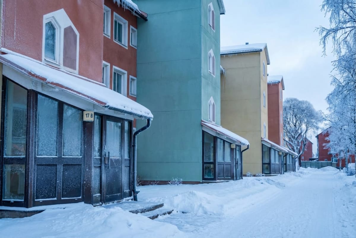 Bilden visar en gata täckt av snö i bostadsområde under vintertid. Till vänster syns flera flerfamiljshus i olika färger: roströd, blågrön och gul. Husen har vita fönster och några synliga stuprör. Husfasaderna är täckta av snö och is, och istappar hänger från taken på flera byggnader. Framför byggnaderna finns snötäckta farstukvistar med nummer, till exempel "17 B" syns tydligt på en av dem. Marken är helt täckt av ett tjockt snötäcke, och en upplogad gångväg eller körbana löper framför husen. Till höger i bilden står snötäckta träd med bara grenar vid sidan av vägen. Himlen är delvis molnig och ljus, vilket indikerar dagtid.