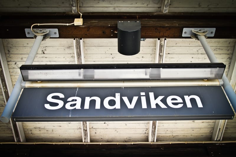 Bilden visar en skylt med texten 'Sandviken' i vit text på en mörkblå eller grå bakgrund. Skylten är upphängd under ett tak av träbalkar och ljusa träpaneler. Ovanför skylten sitter ett avlångt lysrörsarmatur och en svart, cylindrisk högtalare. Elekt
