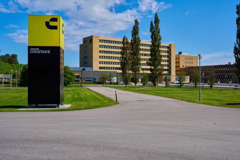 Bilden visar en utomhusmiljö med en stor gul och svart skylt i förgrunden, märkt med texten "SANDVIK COROMANT" och en logotyp. Skylten är placerad på en gräsmatta. Bakom skylten och gräsmattan slingrar sig en asfalterad väg med en uppfart. I bakgrund
