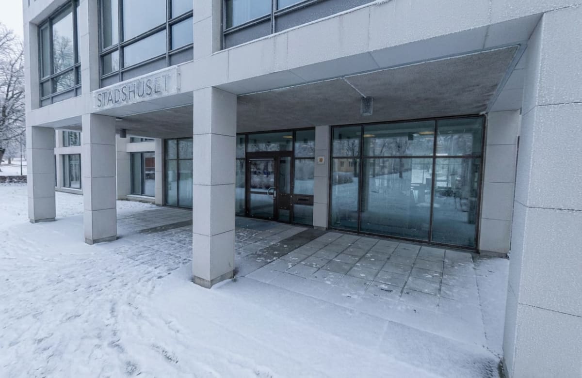 Bilden visar entréområdet till en byggnad som heter "STADSHUSET". Byggnaden är modern med stora fönster och en grå fasad. Entrén har ett utskjutande tak som stöds av flera kvadratiska pelare. Markytan framför entrén är delvis täckt av snö, medan en del av gångvägen närmast byggnaden är av stenplattor som är mindre snötäckta. Det finns spår i snön. Fönsterrutorna på byggnaden reflekterar omgivningen, inklusive träd som också är täckta av snö eller frost, vilket tyder på kallt väder. En dörr med en handtag och en brevlåda syns i mitten av entrén. Byggnaden och dess omgivning är täckta av ett tunt lager frost eller snö som ger en ljus framtoning.