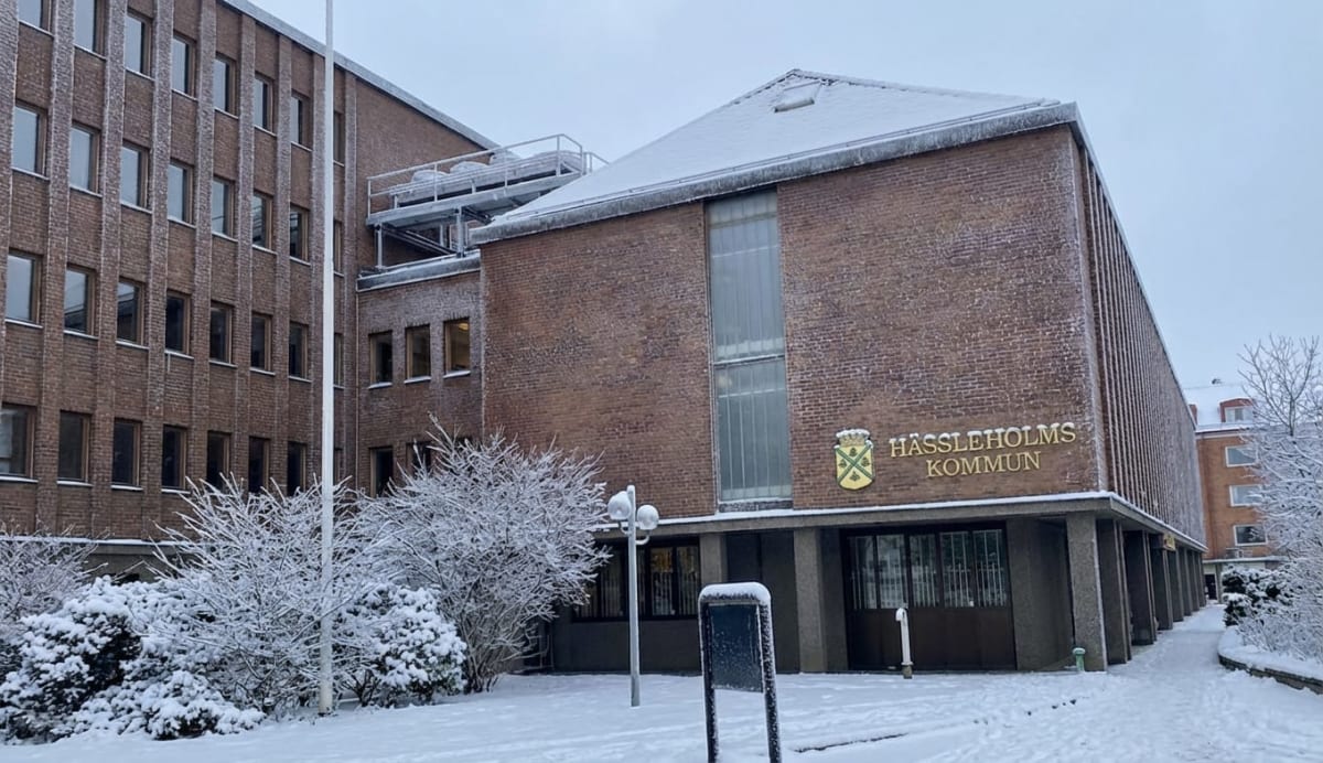 Bilden visar fasaden till en större tegelbyggnad täckt med snö på taken och på marken framför. Hela markytan i förgrunden är snötäckt, och flera buskar och träd syns med snö på grenarna. Fasaden är huvudsakligen av rött tegel med en ljusgrå grund. Ett skyltfönster eller en stor fönstersektion i mitten av byggnaden är synlig. Till höger om denna sektion, på fasaden, sitter en skylt med ett emblem och texten "HÄSSLEHOLMS KOMMUN" i guldfärgade bokstäver. Till vänster om byggnaden sträcker sig en annan del av byggnaden med flera rader av fönster. En vit flaggstång står vertikalt i bildens vänstra del. Framför byggnaden syns ett par lampor på en stolpe samt en fristående skylt eller anslagstavla, båda delvis täckta med snö.