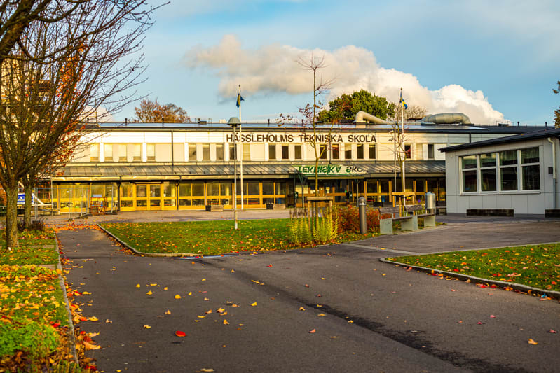 En bild som visar en byggnad med texten "HÄSSLEHOLMS TEKNISKA SKOLA" på fasaden. Framför byggnaden finns ett asfalterat område med löv utspridda, samt gräsmattor med viss växtlighet. Träd med kala grenar syns i förgrunden till vänster och intill bygg