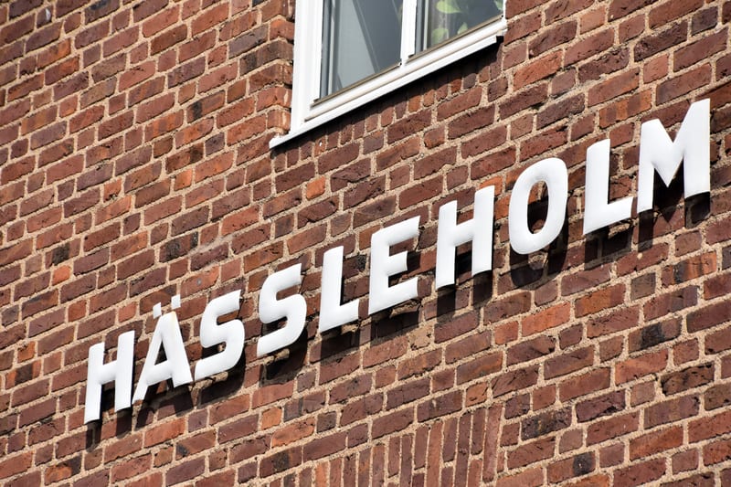 Bilden visar texten "HÄSSLEHOLM" i stora vita versala bokstäver monterade på en röd tegelvägg. Bokstäverna har en skuggverkan från direkt solljus, vilket indikerar att de är tredimensionella eller att belysningen kommer snett bakifrån. Ovanför texten