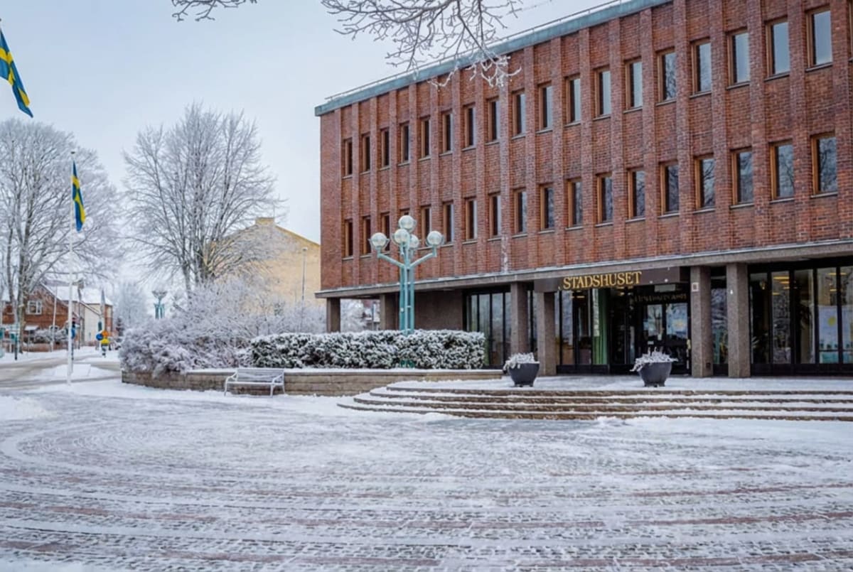 En exteriörbild tagen på dagtid som visar en tegelbyggnad, förmodligen ett stadshus, i ett vinterlandskap. Byggnaden har flera rader med fönster och en ingång med texten "STADSHUSET" ovanför. Framför byggnaden finns ett snötäckt torg med runda trappsteg som leder upp till entrén. Till vänster i bilden syns snötäckta buskar och träd som är täckta av frostrim. Ett par svenska flaggor på stänger står vid sidan om den snötäckta gatan i bildens vänstra del. Hela marken är täckt av snö och frost. En bänk är placerad bredvid buskarna. Himlen är ljus och något molnig, och det ger ett generellt ljust intryck trots vinteromständigheterna.
