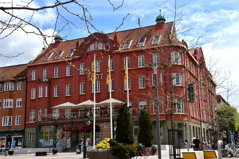 Denna bild visar en röd tegelbyggnad med flera våningar och ett rött tegeltak. Byggnaden har många fönster med vita karmar. På framsidan av byggnaden finns en restaurang eller café med uteservering och vita parasoller. Framför byggnaden syns flaggstä