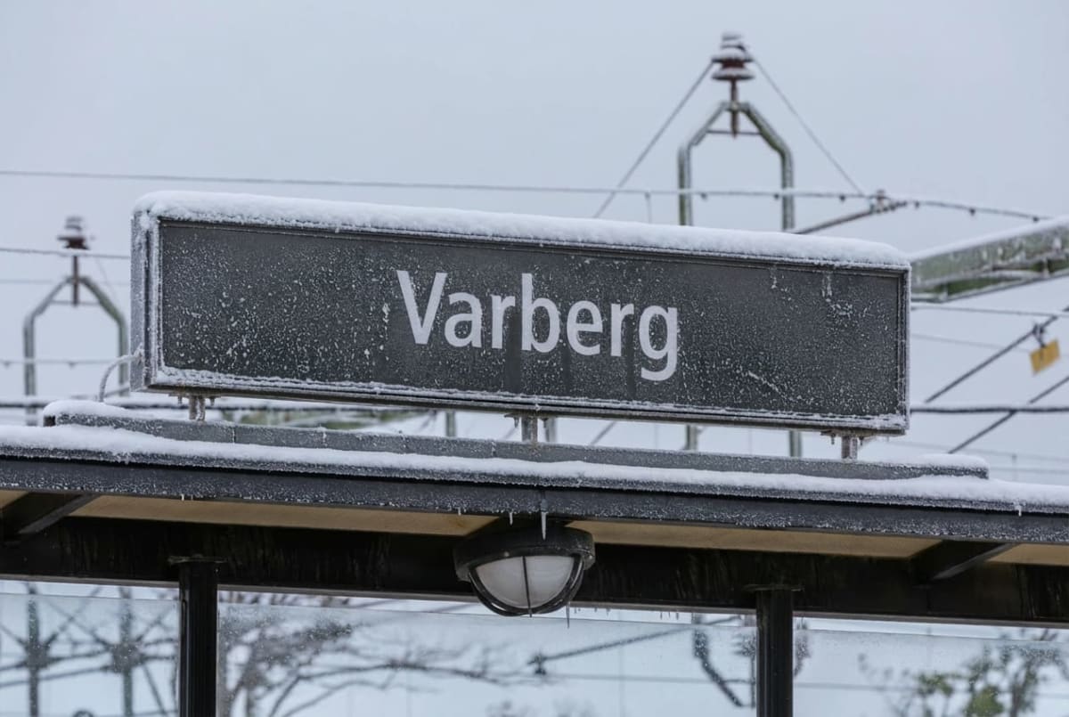 Bilden visar en skylt med texten "Varberg" i vit färg på en mörk bakgrund. Skylten är täckt med snö och rimfrost, vilket indikerar kallt väder. Under skylten syns en del av ett tak med en rund, vit lampa hängande under. Även taket är täckt med snö. I bakgrunden skymtar delar av vad som ser ut att vara kontaktledningar och stolpar för tåg, också de delvis täckta av snö. Hela bilden går i gråa och vita toner.