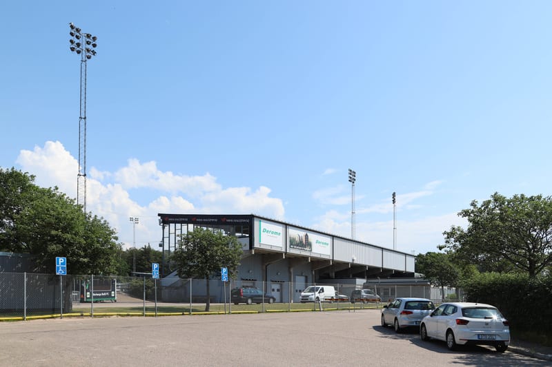 Bilden visar utsidan av en byggnad som liknar en idrottsarena eller sportanläggning under dagtid. En stor grandstand-struktur dominerar bakgrunden med text och logotyper som "Derome" och "Västra Götaland" synliga på banderoller. Framför grandstanden