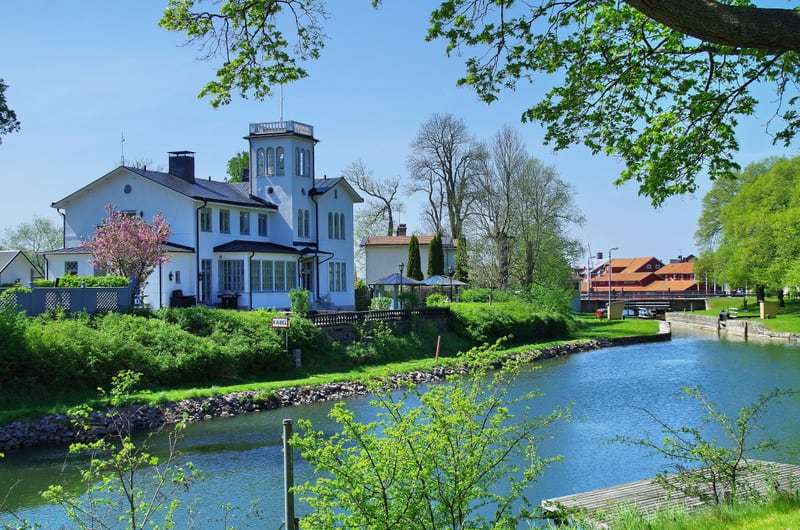 En bild tagen utomhus under dagtid som visar en flod eller kanal med båtars tilläggsplatser, omgiven av grönskande vegetation och byggnader. Till vänster i bilden står en stor vit byggnad med ett torn och flera fönster. Framför byggnaden finns en trä